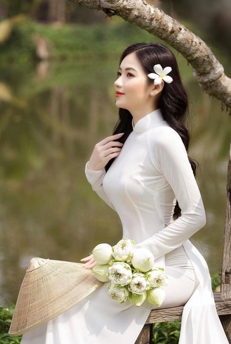 Gái xinh áo dài trắng ngồi trên ghế gỗ cầm hoa sen và nón lá