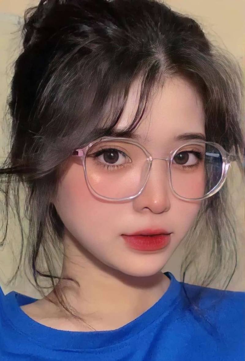 Ảnh gái xinh tóc dài đeo kính mặc áo thun xanh trước bức tường trơn