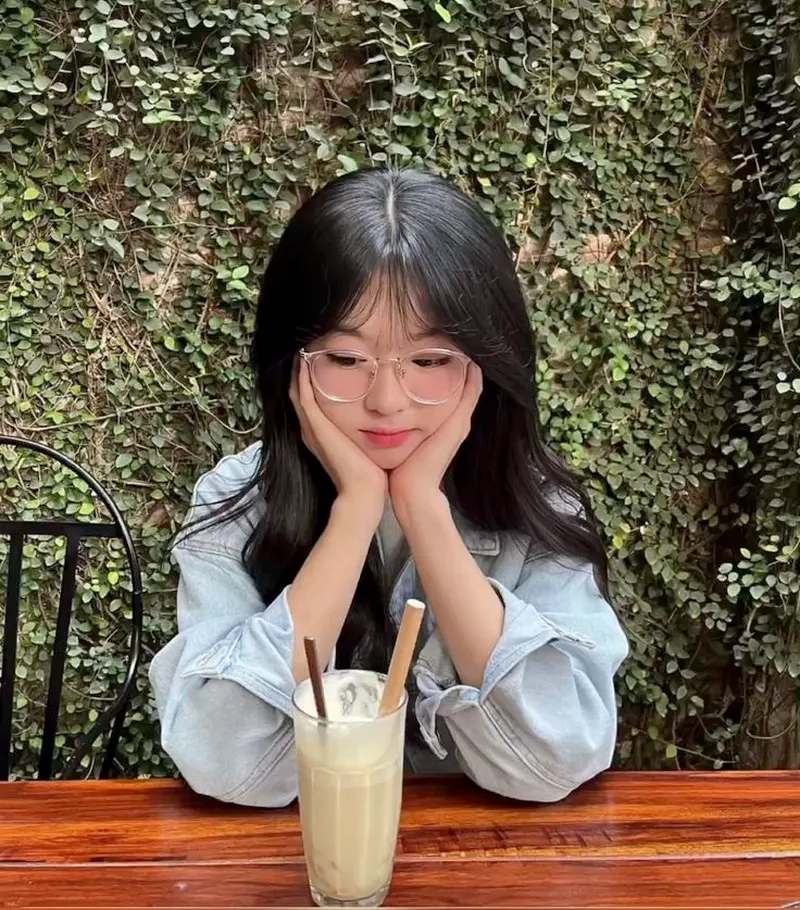 Ảnh gái xinh tóc dài đeo kính hai tay chống cằm ngồi uống sinh tố