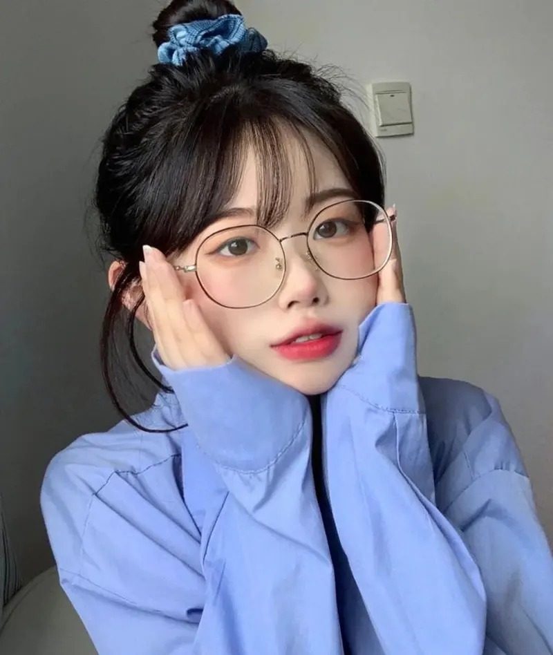 Ảnh gái xinh tóc dài đeo kính mặc áo sơ mi xanh trước bức tường trơn