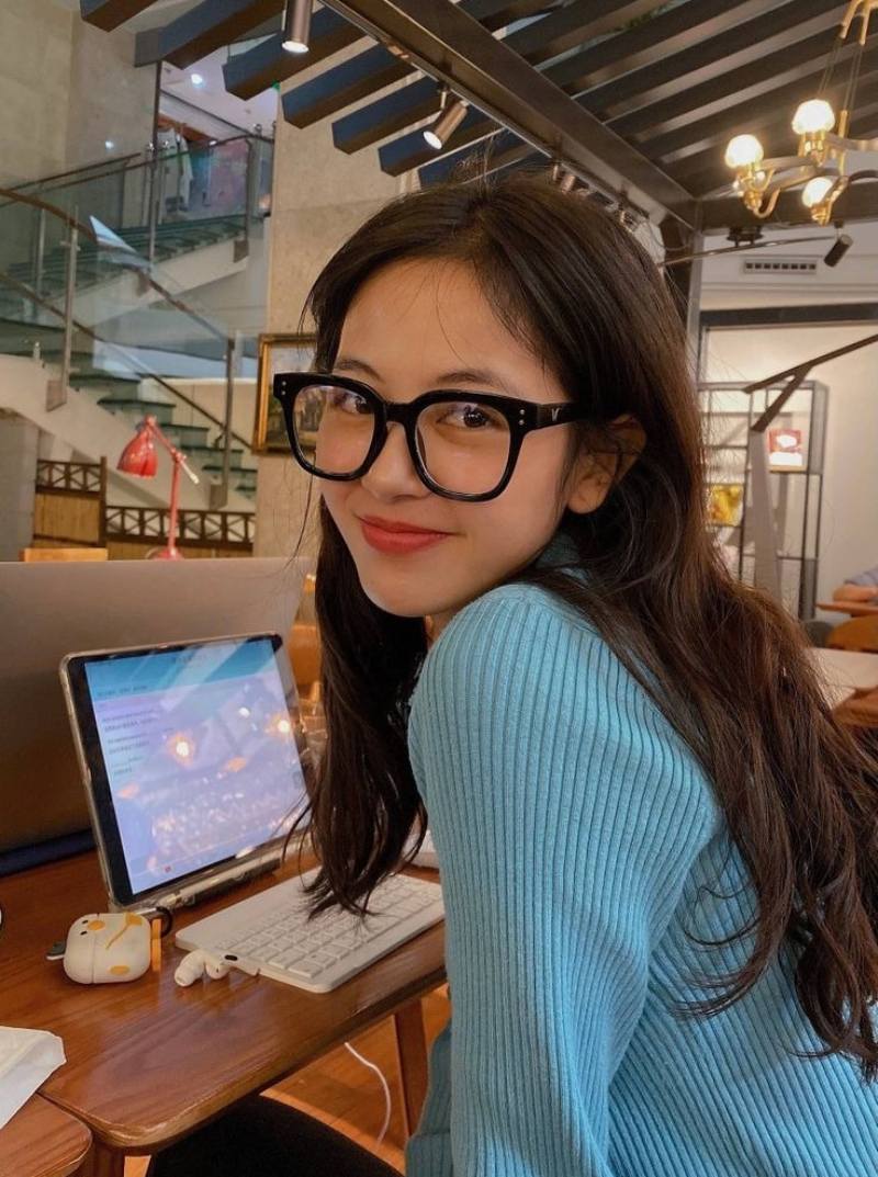 Ảnh gái xinh tóc dài đeo kính cười nhẹ, ngồi làm việc với máy tính